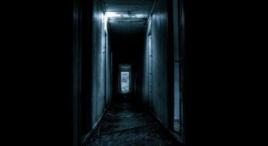 Photograph of a very dark hallway with one sharp light, making it incredibly eerie