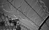 Greyscale image of a barbed wire fence in front of a security camera pointing to the bottom left.