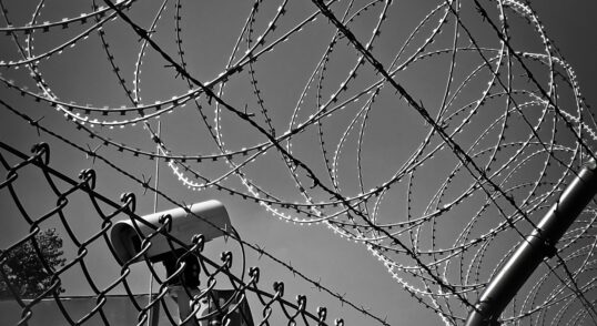 Greyscale image of a barbed wire fence in front of a security camera pointing to the bottom left.