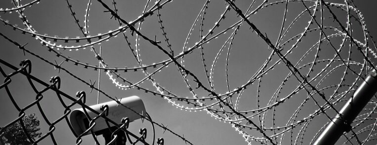Greyscale image of a barbed wire fence in front of a security camera pointing to the bottom left.